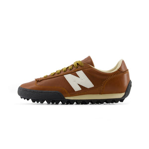 Lateral side profile of Donald Glover x New Balance RC30 "Sea Salt / Macadamia Nut" shoes, featuring a smooth cognac brown leather upper with contrast stitching, a white leather N logo, mustard green laces, off-white midsole, and a black rippled studded rubber outsole.
