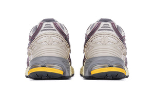 Rear heel view of a pair of grey and purple New Balance shoes 1906R model, featuring light beige molded TPU support cages with square NB branding, metallic mauve trim accents, off-white mesh collars, grey padded lining, and cream N-ergy cushioned midsoles with vibrant orange heel pods.