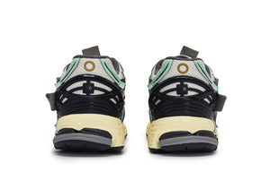 Rear heel view of a pair of grey, green, and blue New Balance shoes 1906A model, featuring black TPU heel support cages with NB branding, mint green accents, circular heel motifs, grey mesh collars, and aged yellow N-ergy cushioned midsoles.