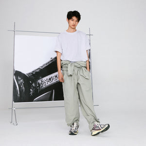 Studio full-body editorial lifestyle shot of a model wearing grey and purple New Balance shoes 1906R, paired with a white t-shirt and loose sage green trousers tied at the waist, standing in front of a framed black-and-white art backdrop.