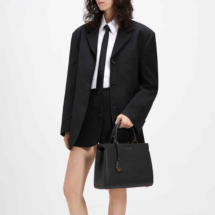 CHARLES&KEITH Mirabel top handle bag in black with gold-tone accents, carried by a model in a black suit.