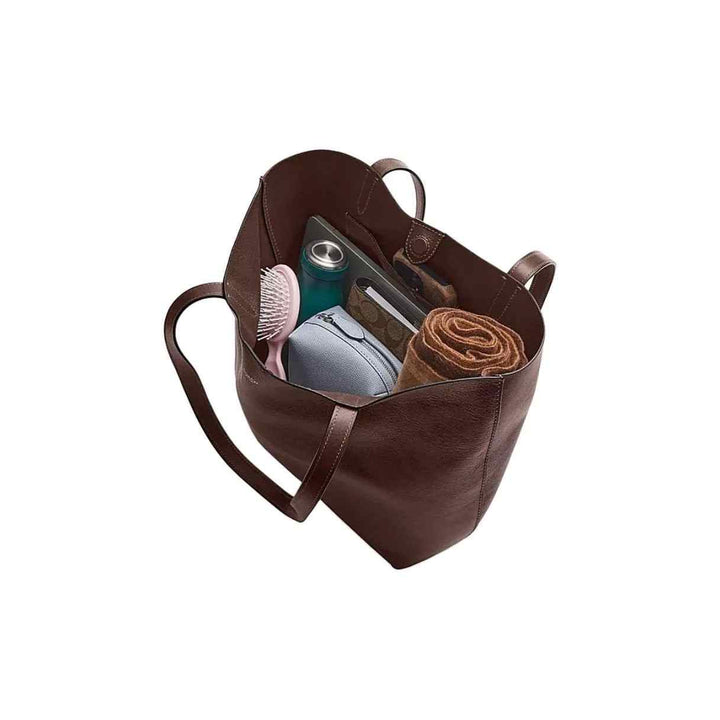 brown leather Manhattan tote handbag by Coach filled with daily essentials including a hairbrush, water bottle, cosmetics, and a rolled towel