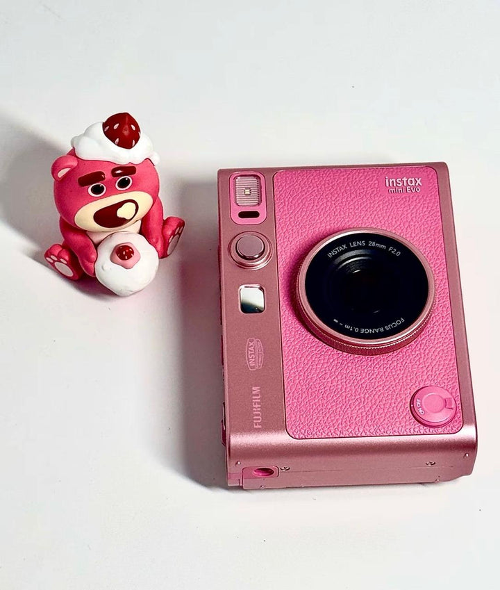 كاميرا فورية وردية FUJIFILM Instax Mini Evo مع تصميم أنيق وتقنيات متقدمة، تقع بجانب لعبة وردية صغيرة.