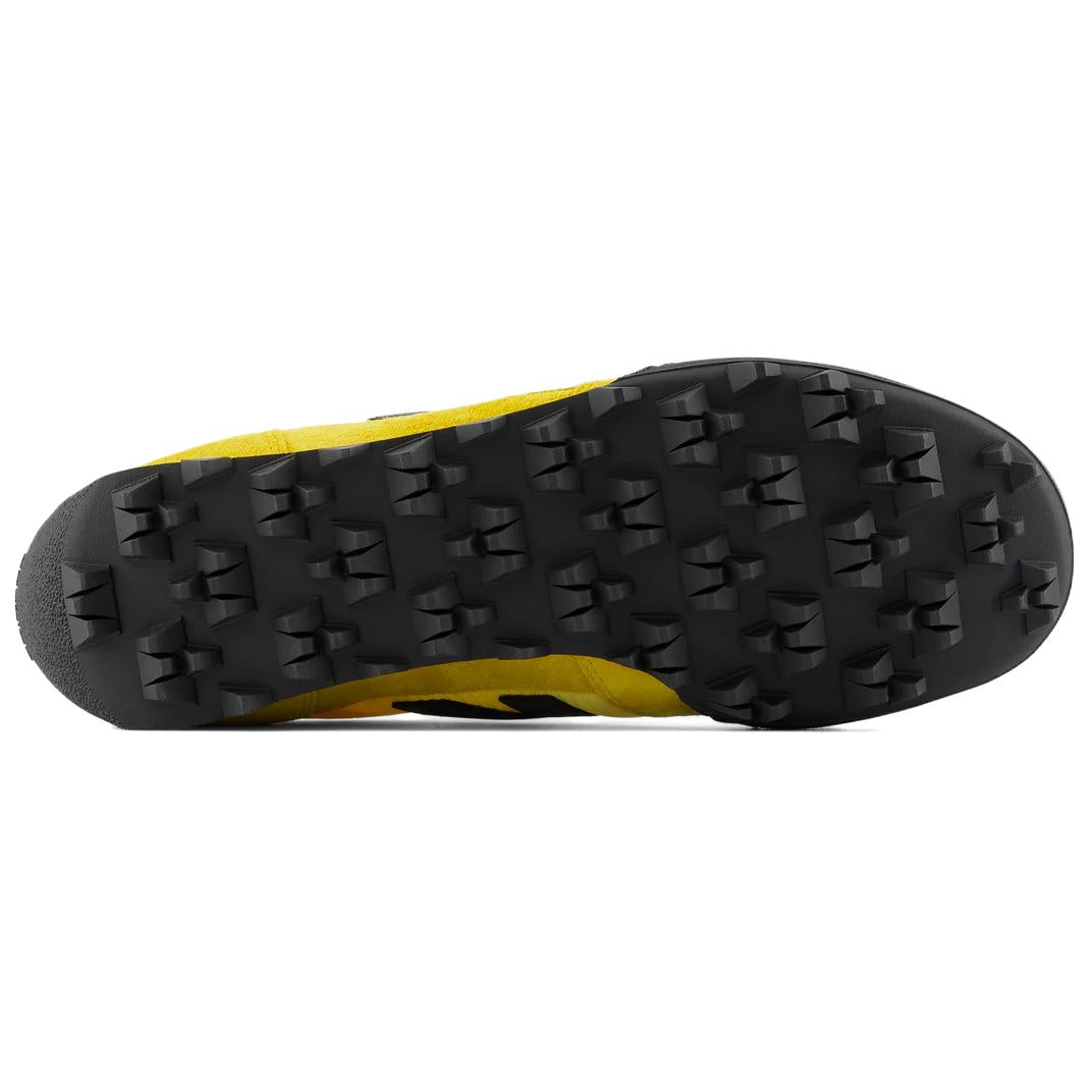 Bottom outsole view of yellow and black New Balance RC30 shoes, featuring a black rubber tread pattern with exaggerated angular studs across the entire sole.