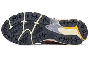 Bottom outsole view of grey and purple New Balance shoes 1906R model, featuring dark grey rubber traction tread pattern, cream flex grooves, bright orange heel highlights, and a grey midfoot Stability Web arch support shank.