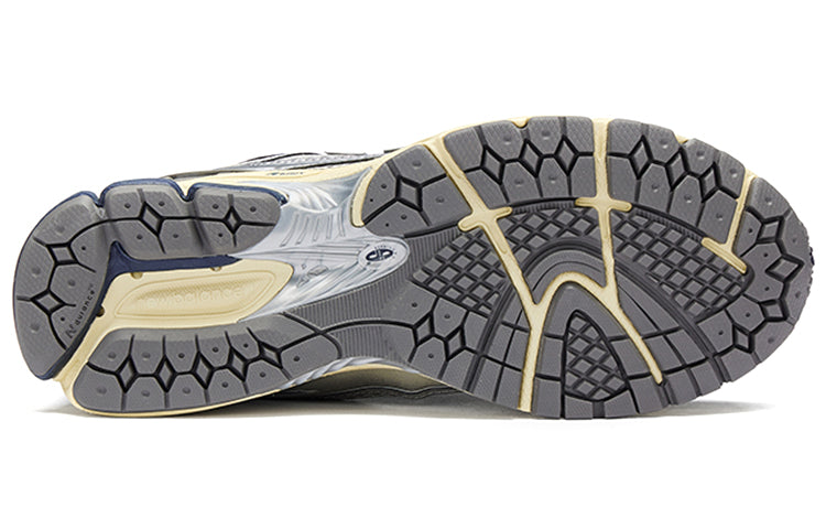 Bottom outsole view of metallic silver and navy blue New Balance shoes 1906R model, featuring dark grey rubber traction tread pattern, cream flex grooves, navy blue heel accents, and a silver midfoot Stability Web arch support shank.