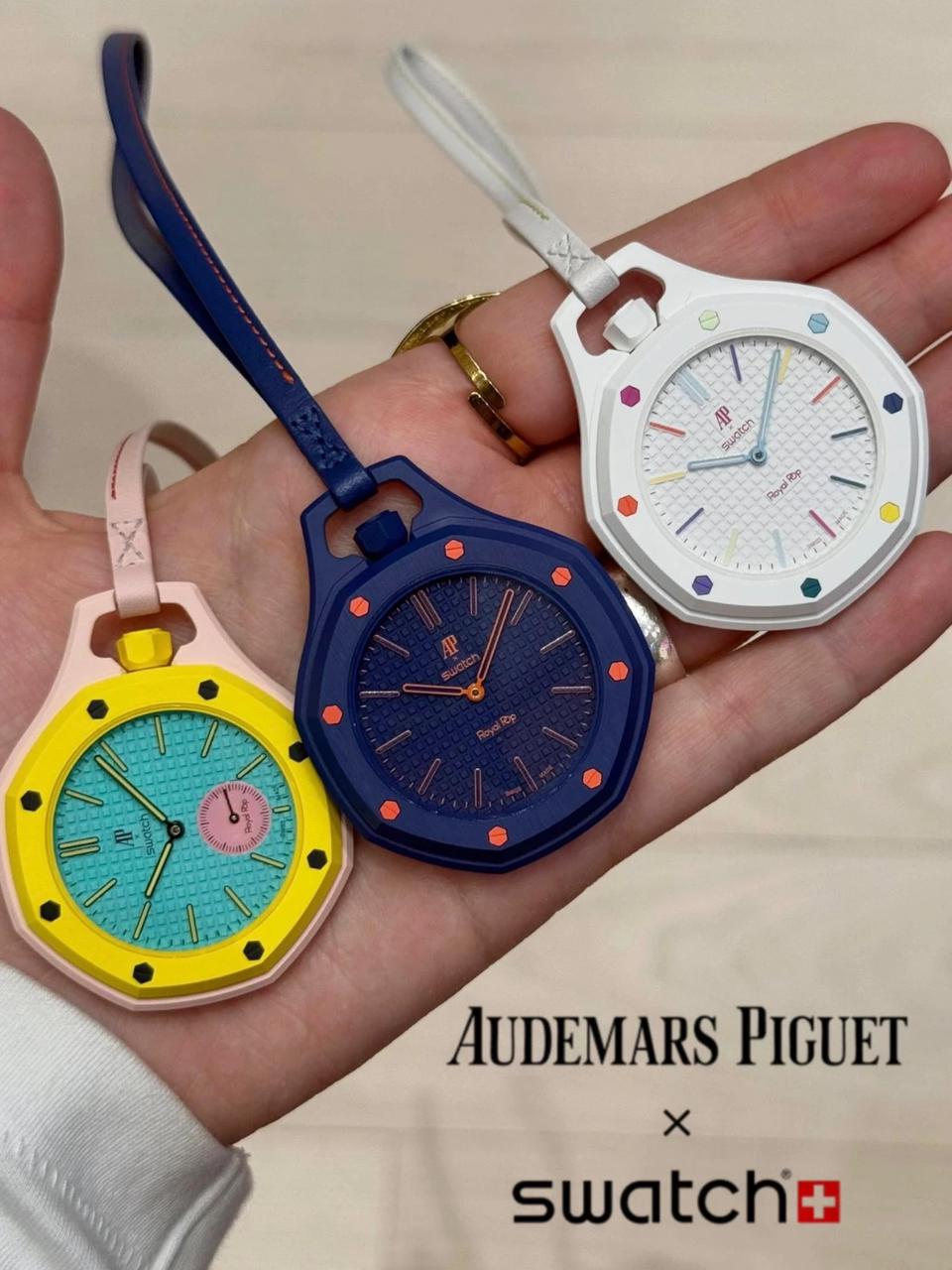 Three Audemars Piguet x Swatch Royal Pop pocket watches in white, dark blue, and yellow with pink accents held in a hand by their leather straps.