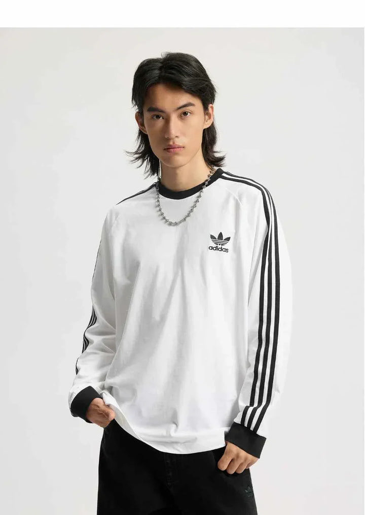 رجل يرتدي adidas تيشيرت قطني كلاسيكي بأكمام طويلة أبيض مع خطوط سوداء وشعار Trefoil على الصدر