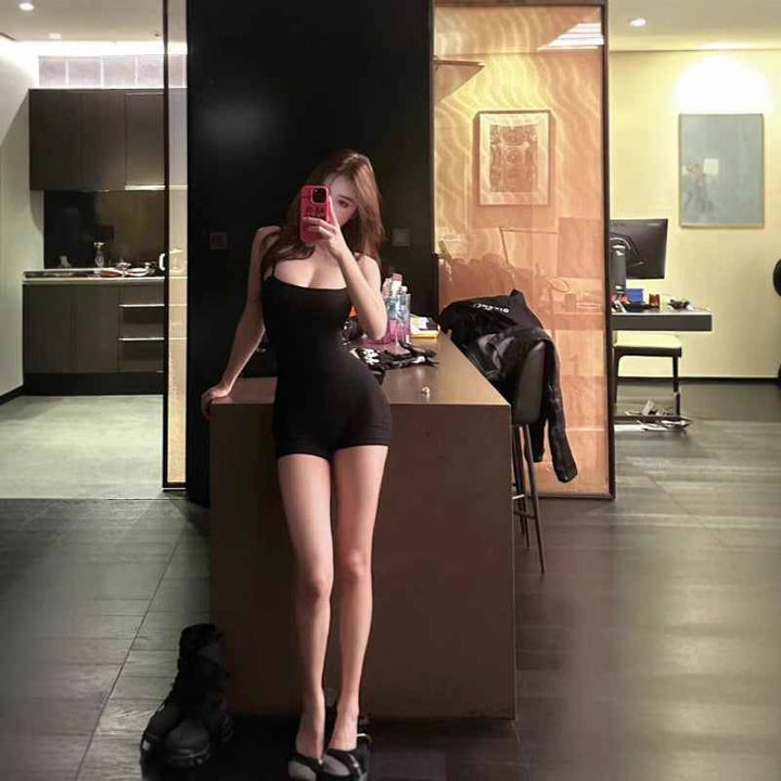 Woman wearing black SKIMS short romper with thin straps taking a mirror selfie in modern room