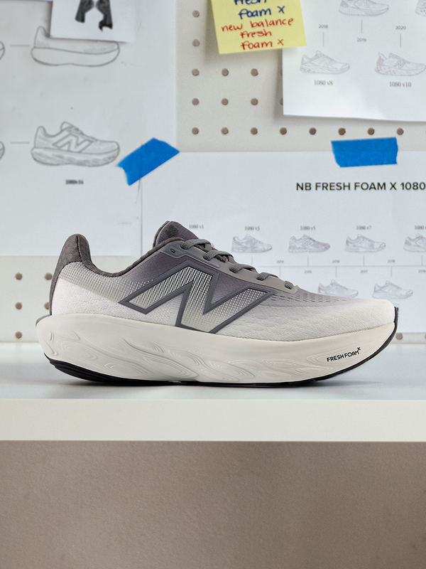 Side view of a gray New Balance Fresh Foam X 1080 v14 running shoe displayed on a white surface with design sketches in the background