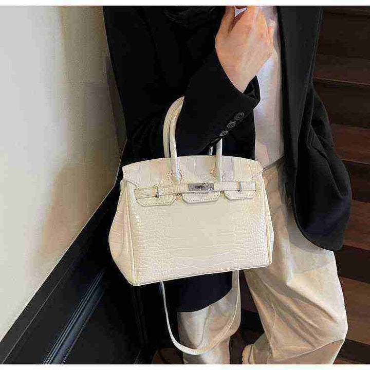 White Birkin-style women's handbag with crocodile texture, elegant and high-quality finish, held by a person wearing a black blazer and beige pants.