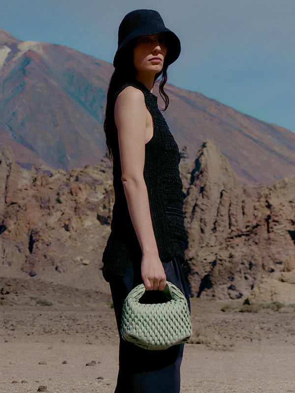 Woman in black outfit and hat holding a handwoven Polène Béeri green leather handbag against a rocky desert landscape