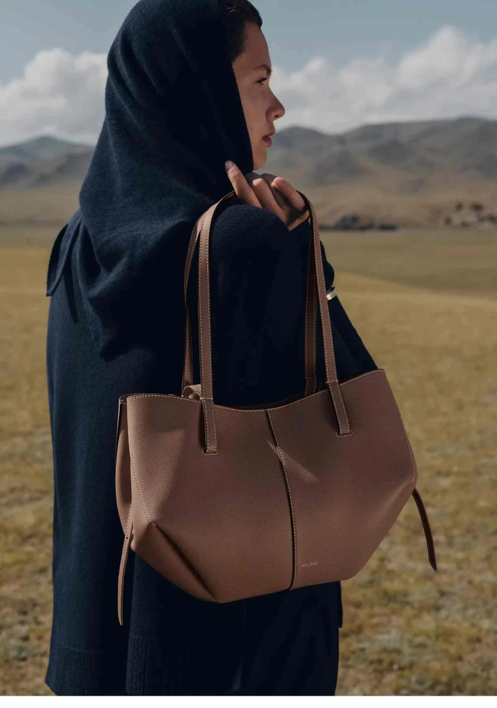 Polène Cyme Mini tote bag in maroon leather with soft curves, worn by a woman outdoors against scenic hills.