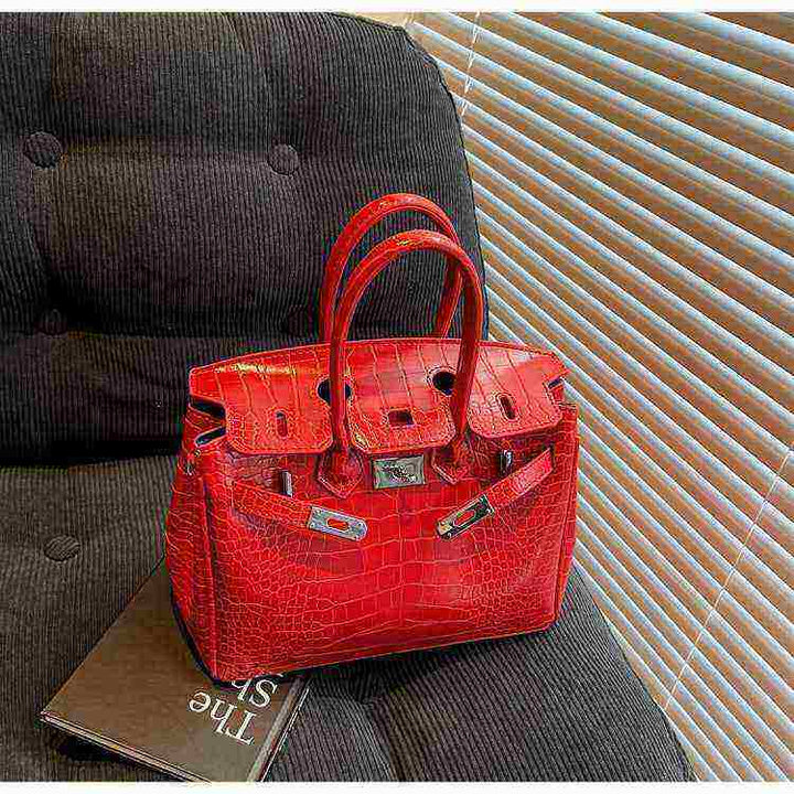Red women's Birkin-style handbag with crocodile pattern and silver hardware placed on a dark chair.