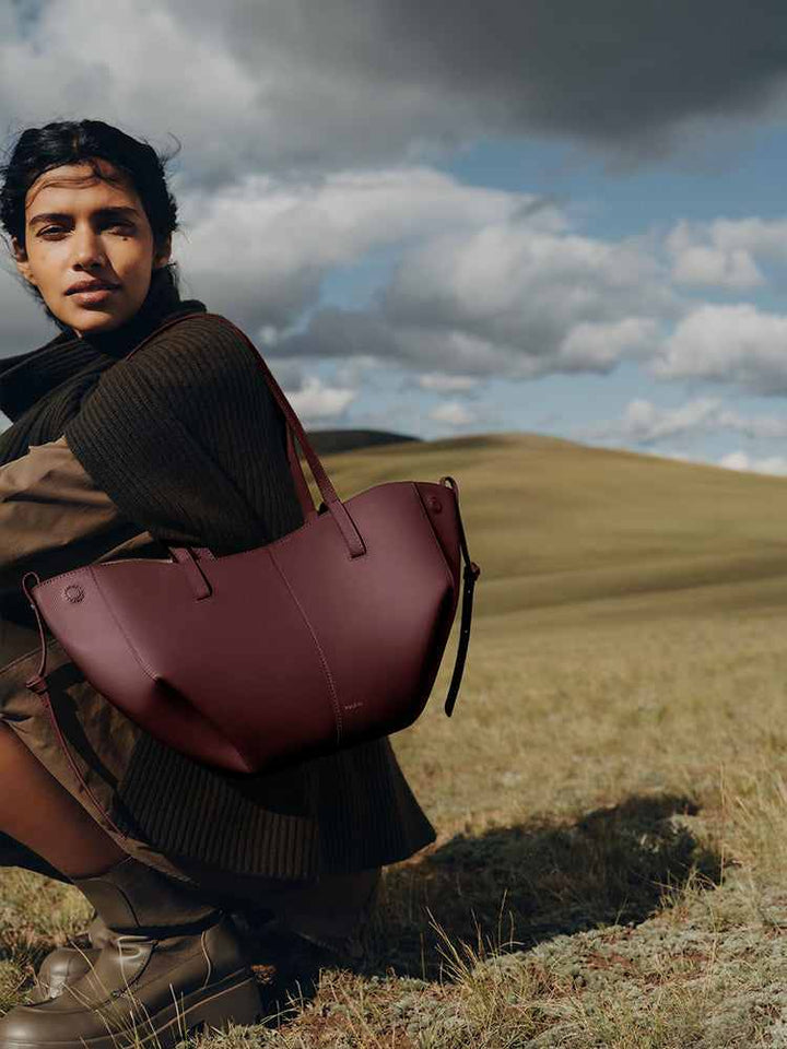Polène Cyme Mini maroon leather tote bag with soft curves worn by a woman outdoors in a natural landscape