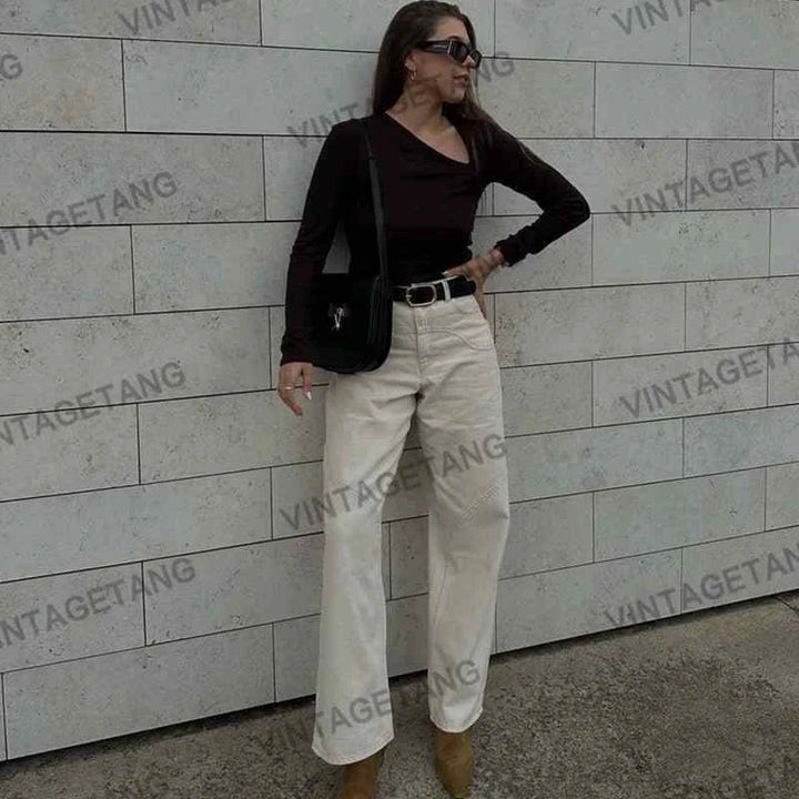 woman wearing high-waisted beige wide cotton pants with black long sleeve top and black shoulder bag standing against tiled wall