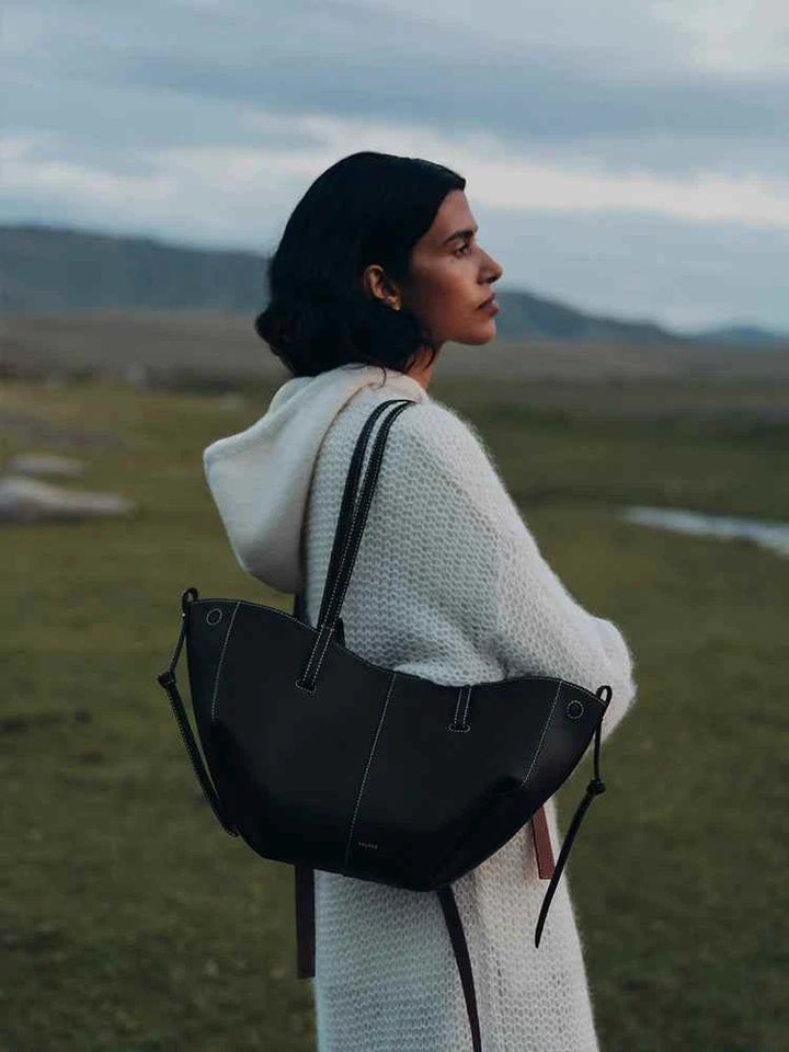 Polène Cyme Mini black leather tote bag with smooth curves and dual carrying straps worn by woman outdoors