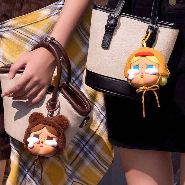 Two Crybaby plush keychains featuring crying faces with large white tears, one wearing a brown bear hat and the other a yellow duck hat, attached to handbags.