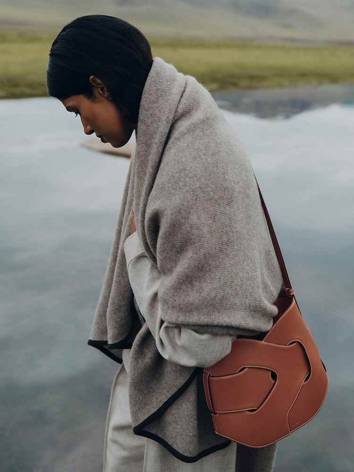 Woman wearing gray shawl and carrying cognac leather Polène Nodde Hobo bag outdoors near water