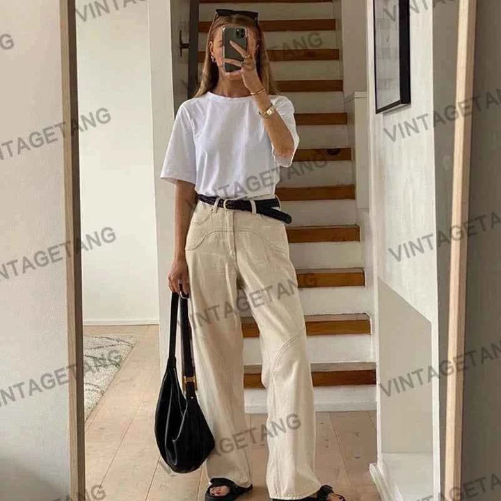 Woman wearing beige high-waisted wide cotton pants with a white t-shirt and black sandals, holding a black bag in a modern interior.