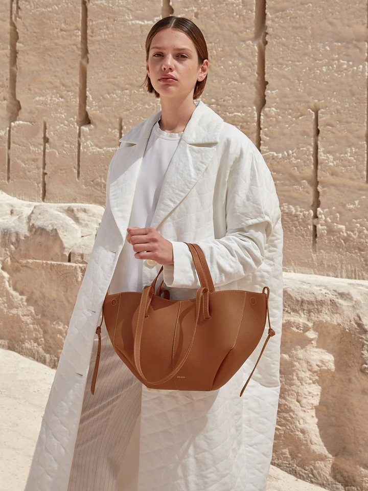 Woman wearing white coat holding camel Polène Cyme Mini leather tote bag with soft curves and dual straps.