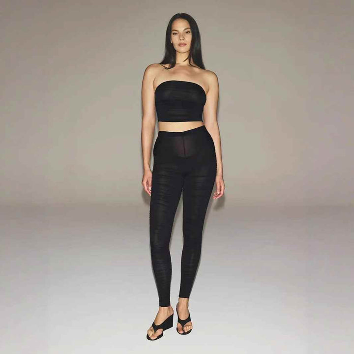 Woman wearing black SKIMS ruched bandeau crop top and matching leggings standing against plain background