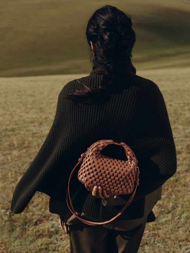 Woman holding a woven brown Polène Béeri leather handbag with top handle and adjustable shoulder strap, standing outdoors in a natural setting