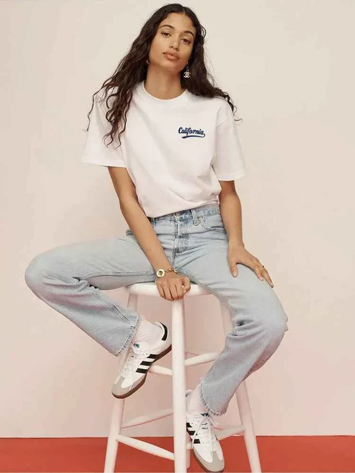 Woman wearing white تيشيرتات California Club with blue California print and light blue jeans sitting on white stool