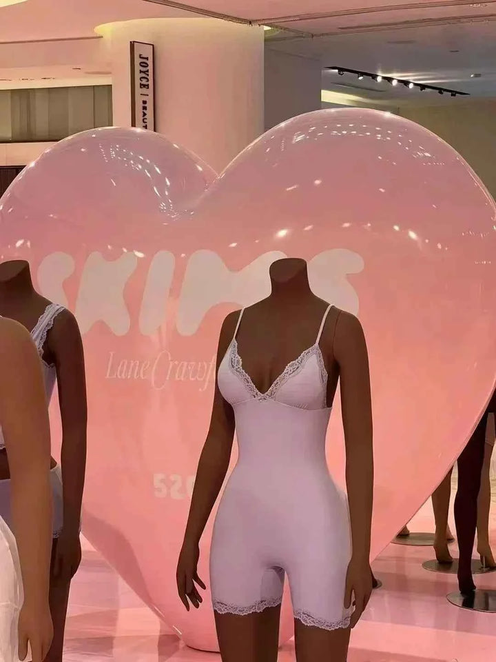 mannequin wearing light pink lace trimmed short-sleeve bodysuit in front of pink heart-shaped backdrop with SKIMS logo
