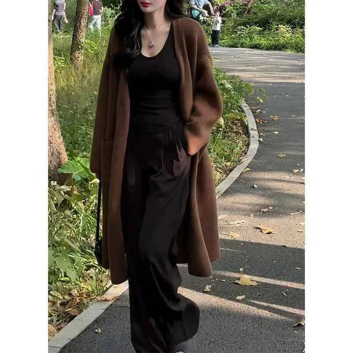 Woman wearing dark brown long wide winter cardigan over black outfit walking outdoors on a pathway surrounded by greenery