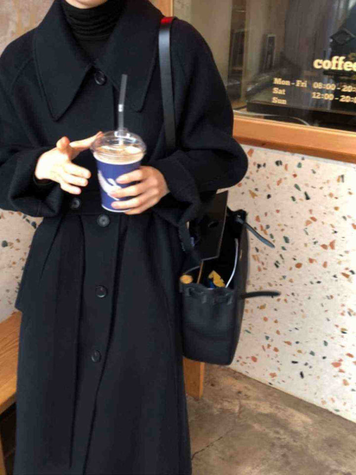 black long winter coat made from luxurious goat wool with button closure and belt detail, styled with a black turtleneck and handbag, worn by woman holding a coffee cup indoors