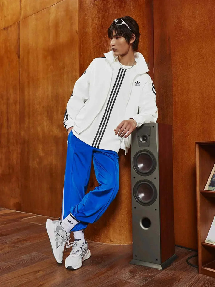 رجل يرتدي Adidas | أديداس جاكيت رياضي كلاسيكي أبيض مع خطوط ثلاثية ويقف بجانب سماعة مكبر صوت