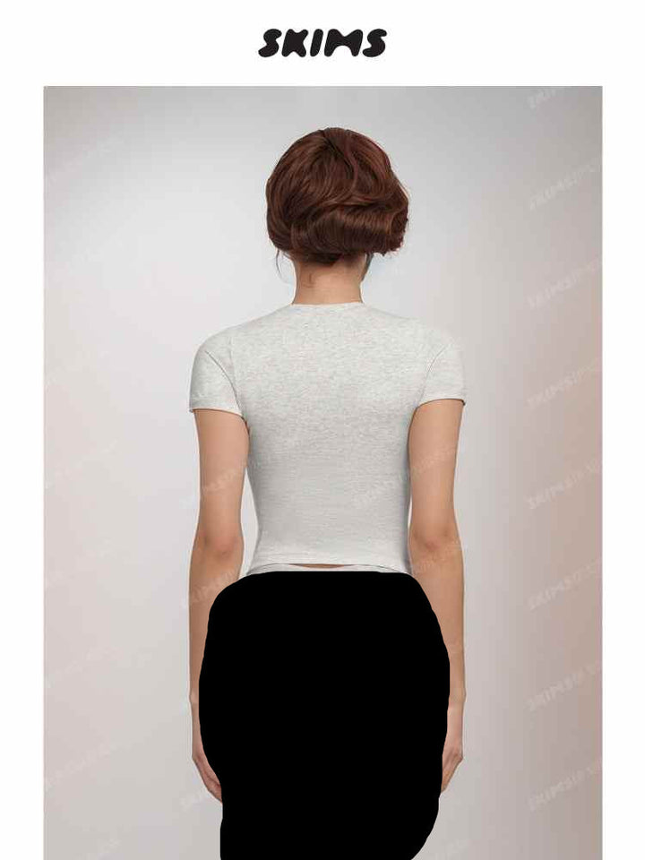 Back view of a woman wearing a fitted light gray SKIMS تيشيرت سكيمز عالمي T-shirt with short sleeves and round neckline.