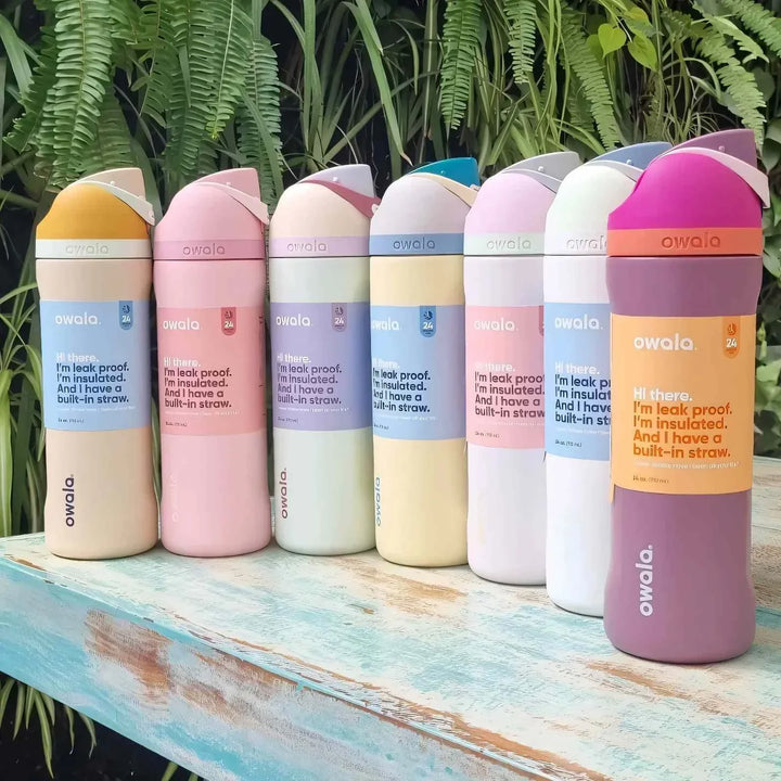 Collection of colorful Owala FreeSip stainless steel insulated water bottles with dual spout lids arranged on wooden surface outdoors