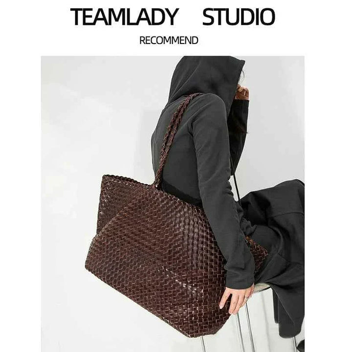 Large dark brown woven handmade natural leather tote bag on shoulder of person in black hoodie and pants sitting on chair