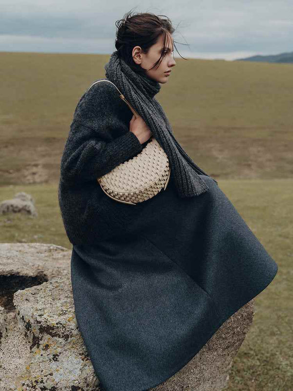 Woman sitting outdoors wearing dark clothing and holding a woven Polène Numéro Dix crescent handbag