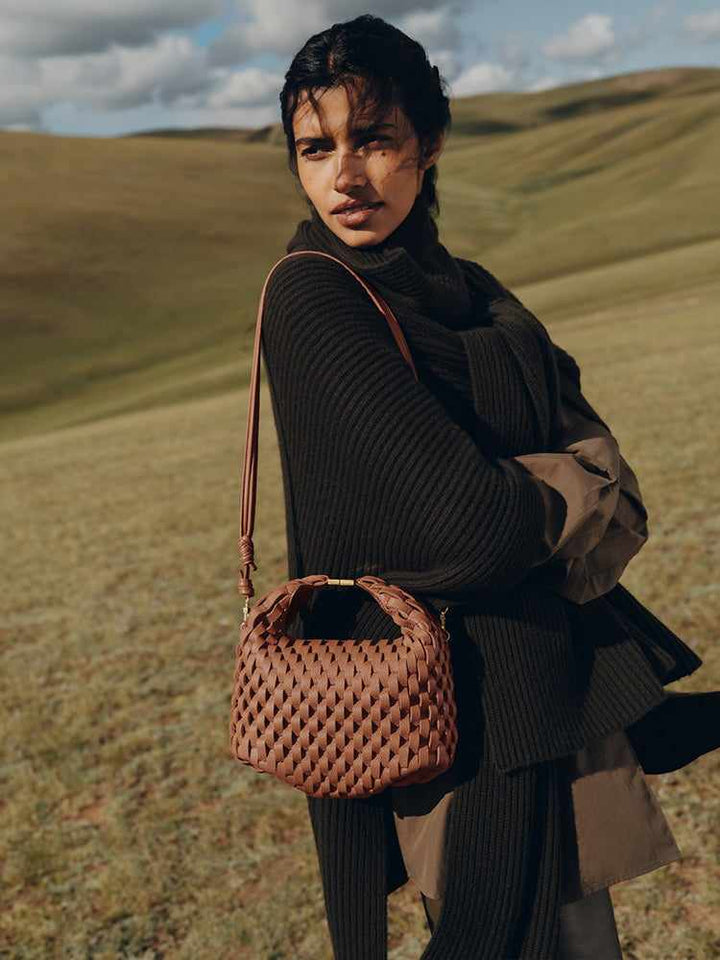 Woman wearing dark outfit carrying woven leather Polène Béeri handbag in cognac color outdoors in a grassy landscape