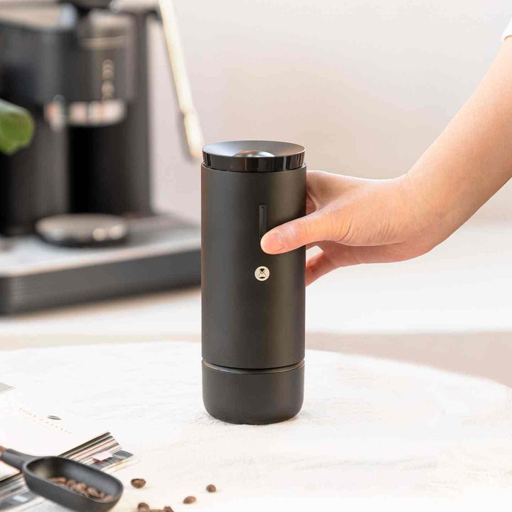 Portable black TIMEMORE WHIRLY 01S electric coffee grinder held by hand on white surface with coffee beans nearby
