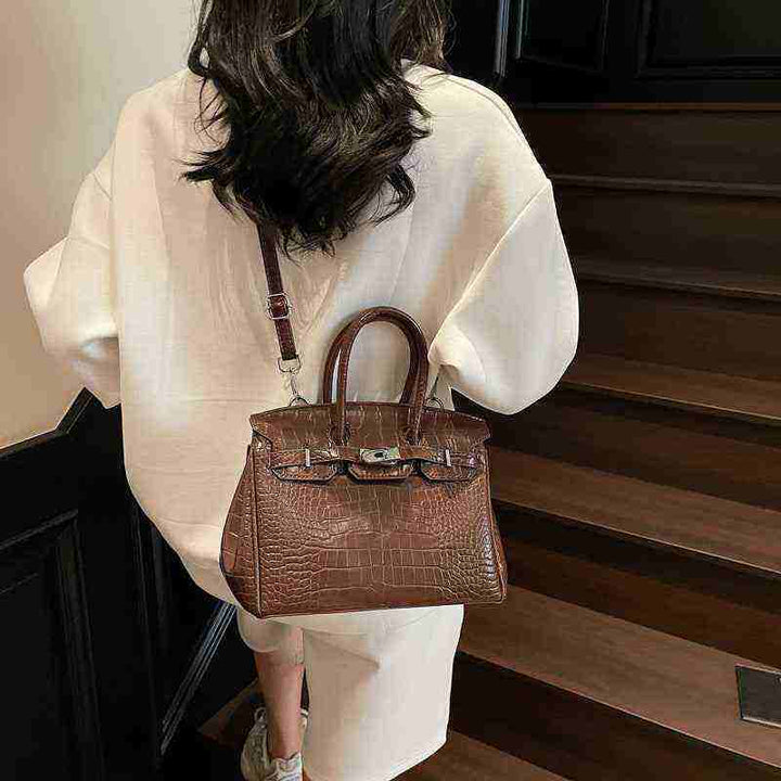 Brown women's Birkin-style handbag with crocodile texture, high-quality leather, and adjustable shoulder strap, worn by a woman in a white outfit on stairs.