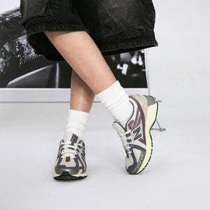 On-foot lifestyle shot of grey and purple New Balance shoes 1906R model, featuring off-white mesh uppers, metallic mauve and slate grey overlays, cream midsoles, and vibrant orange heel pods, paired with slouched white ribbed crew socks and black denim shorts.