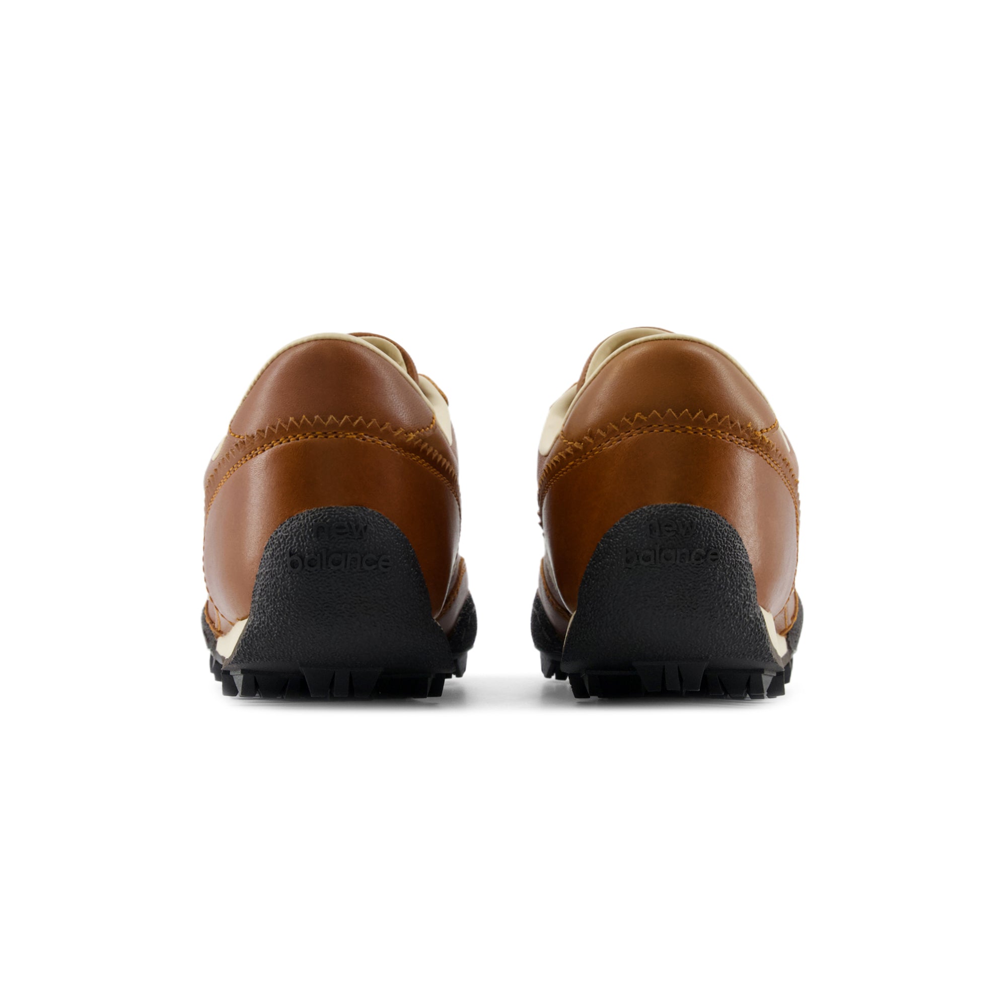 Rear heel view of a pair of Donald Glover x New Balance RC30 shoes, featuring smooth cognac brown leather heels with yellow contrast stitching, serrated collar trim, and an extended black rubber outsole wrap stamped with "new balance" text branding.