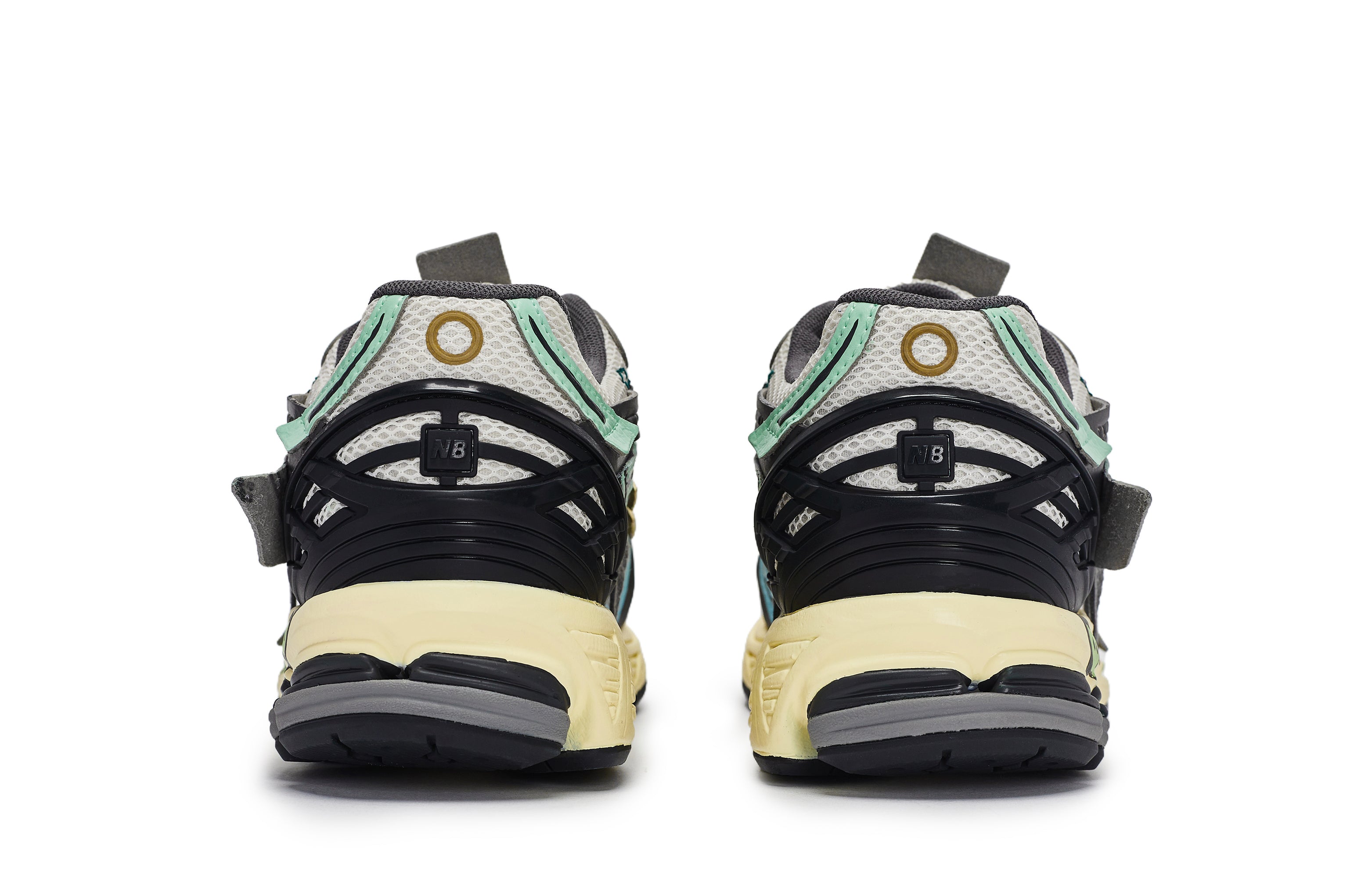 Rear heel view of a pair of grey, green, and blue New Balance shoes 1906A model, featuring black TPU heel support cages with NB branding, mint green accents, circular heel motifs, grey mesh collars, and aged yellow N-ergy cushioned midsoles.
