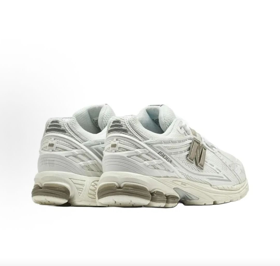 Rear three-quarter view of a pair of cream and off-white New Balance shoes 1906R model, featuring molded off-white TPU heel counters, grey mesh heel accents, beige N-lock branding, and off-white N-ergy cushioned midsoles with translucent grey heel pods.