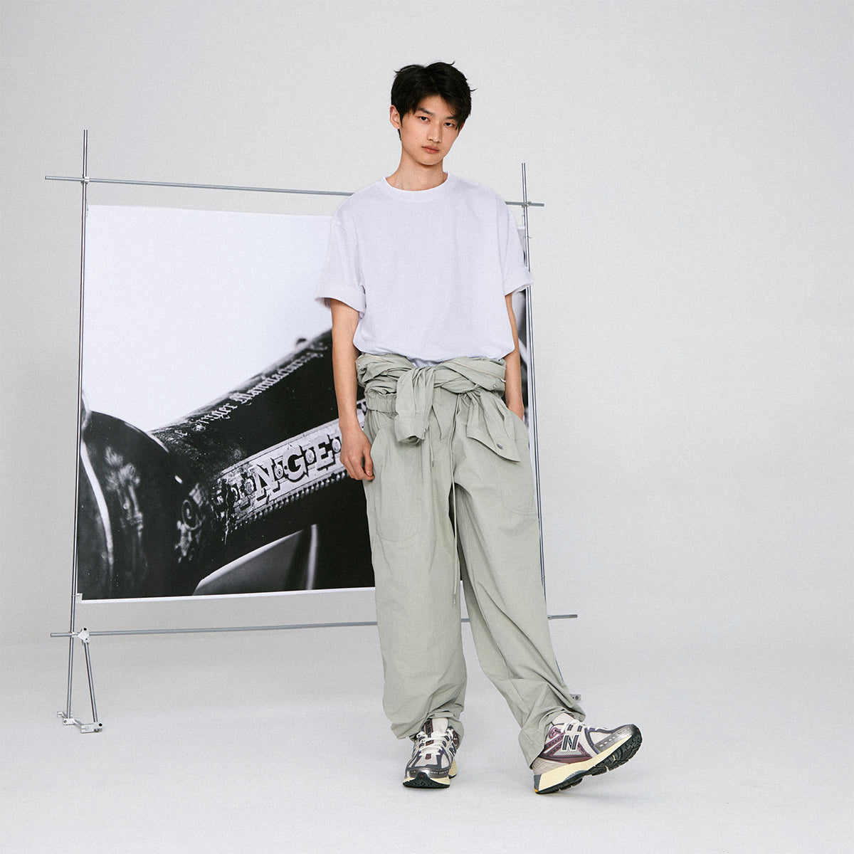 Studio full-body editorial lifestyle shot of a model wearing grey and purple New Balance shoes 1906R, paired with a white t-shirt and loose sage green trousers tied at the waist, standing in front of a framed black-and-white art backdrop.