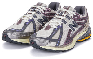 Three-quarter profile shot of a pair of grey and purple New Balance shoes 1906R model, featuring off-white mesh uppers, metallic mauve and slate grey overlays, cream laces, dark grey collar lining, and cream N-ergy cushioned midsoles with orange heel pods.
