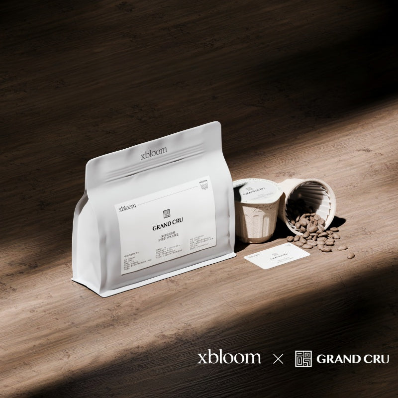 A white bag of XBloom coffee and individual XBloom xpods coffee capsules with loose roasted beans on a wooden surface, showing the 'xbloom x GRAND CRU' collaboration logo.