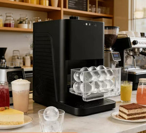 A premium black ice maker machine on a kitchen counter surrounded by iced drinks and desserts, displaying clear crystal ice spheres in its tray.