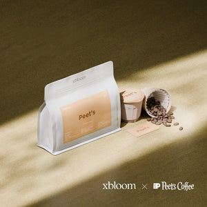 A white bag of Peet's Coffee next to individual XBloom xpods coffee capsules and loose roasted beans on a textured surface, showcasing the 'xbloom x Peet's Coffee' collaboration logo.