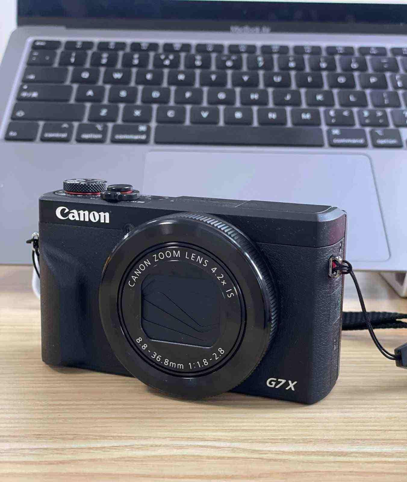Front view of the compact black Canon PowerShot G7 X Mark III sitting on a desk in front of a laptop keyboard.