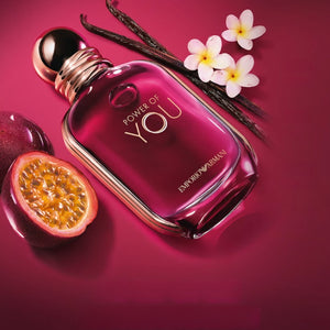 The power of you armani original perfume bottle displayed next to fresh passion fruit slices, vanilla beans, and white blossoms on a deep pink background.